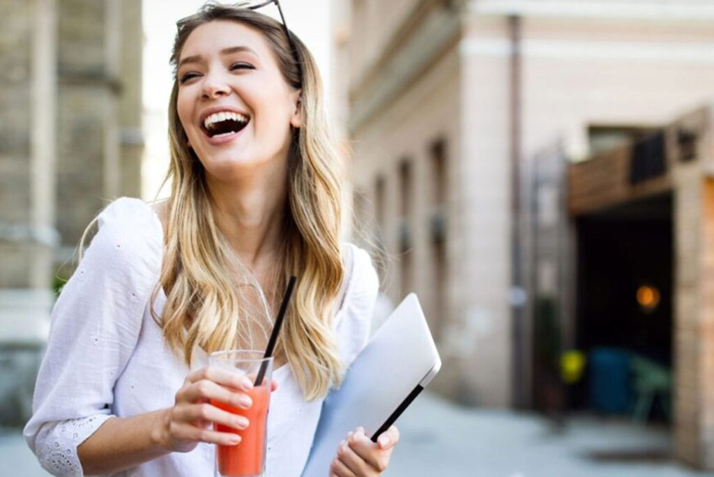 Smiling woman outdoors holding a laptop and drink, representing the power of social media for photographers to gain exposure and bookings.