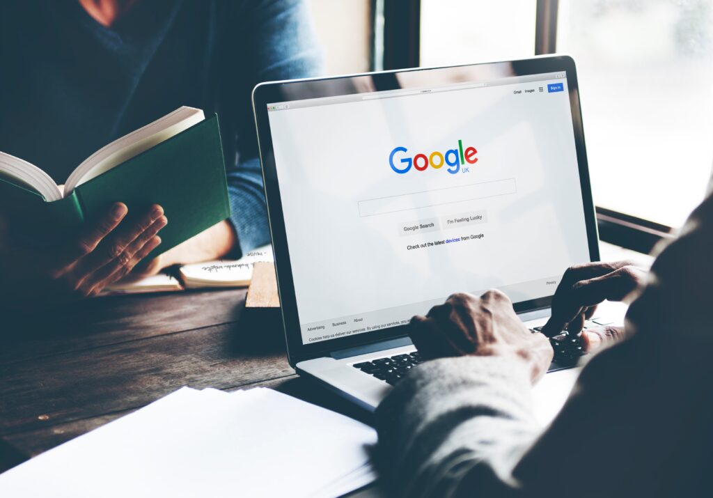 A person using a laptop displaying the Google search page, with another individual reading a book, promoting Google Ads for photographers.