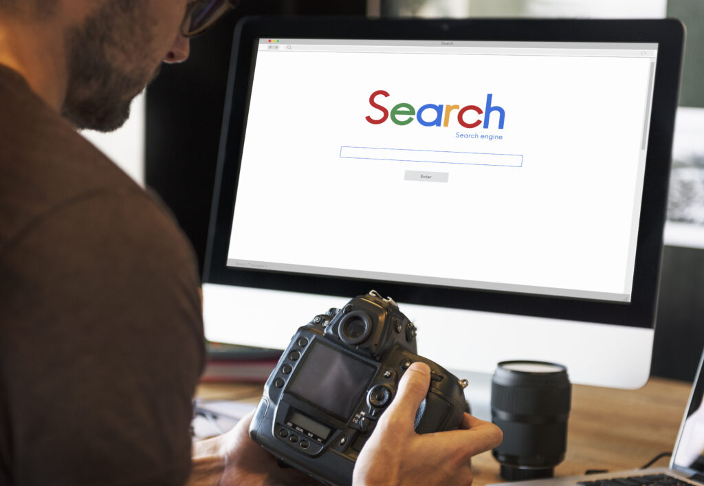 a Photographer Holding a Camera While Working at a Desk with a Monitor Displaying a Search Engine Showcasing Seo Tips for Photographers