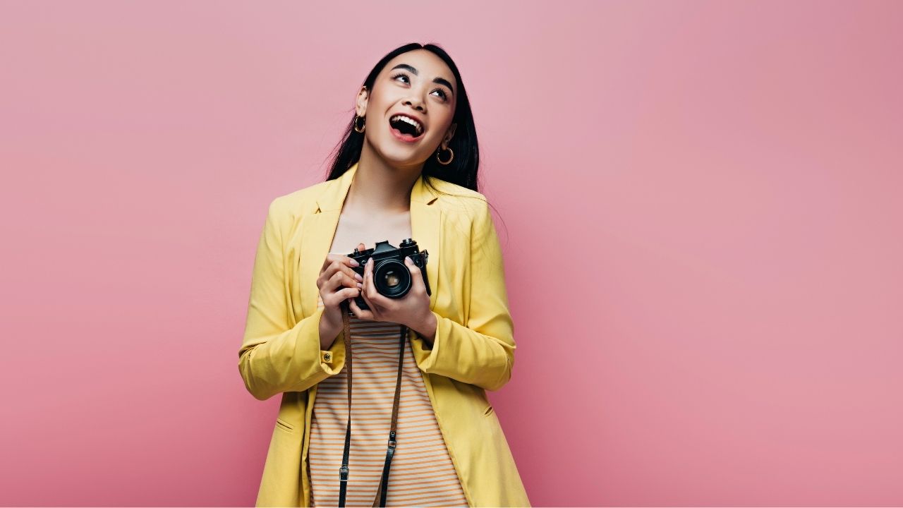 I need to optimize this images for SEO on my photography website. Can you write me the following for this image:  Alternative Text Title Description Please use this key phrase for each and make it sound natural. Key phrase: Backlinks for Photographers The image is being used on this post, so make it relevant to where it is being used:  Smiling female photographer holding a camera against a pink background, representing the power of backlinks for photographers to increase online authority.