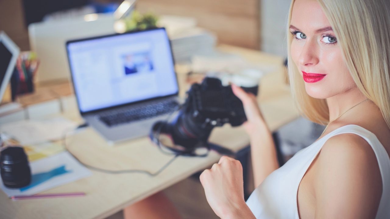 Female photographer holding a camera while working at a laptop, representing research on the best CRM for photographers.