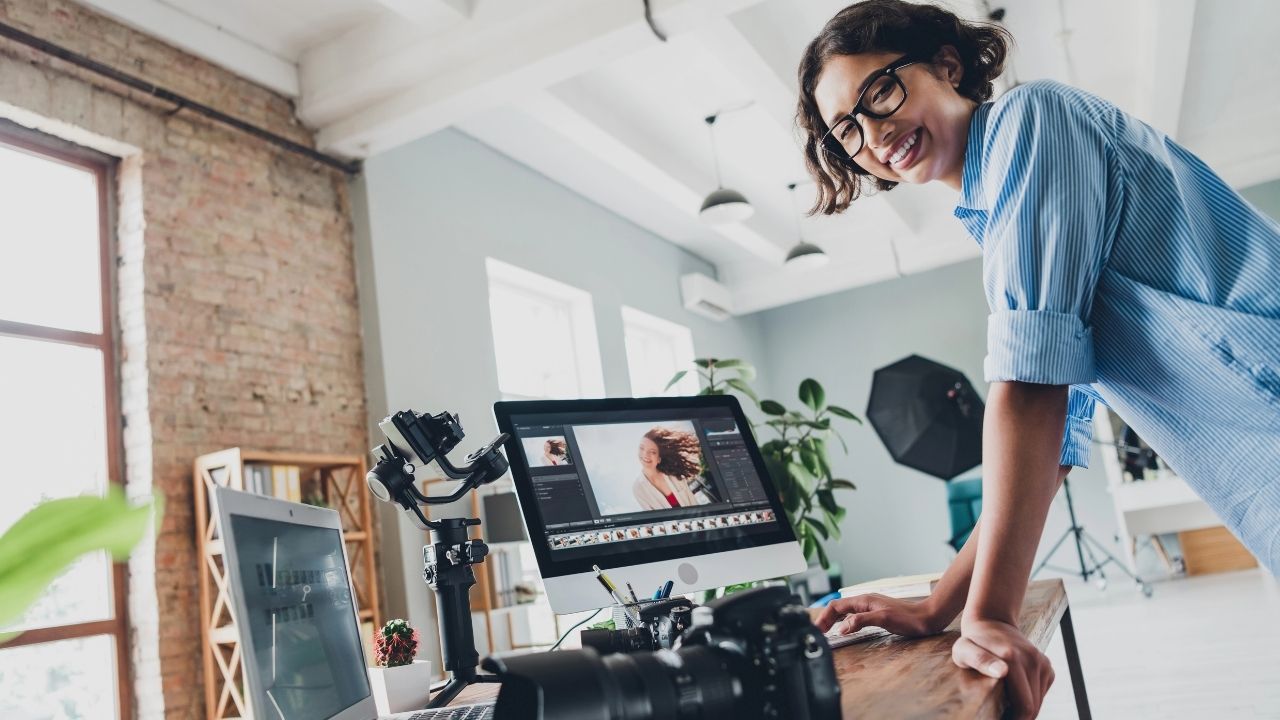 Smiling photographer working at a desk with camera gear and editing software on a computer screen, symbolizing how to design a high-converting photographer homepage.