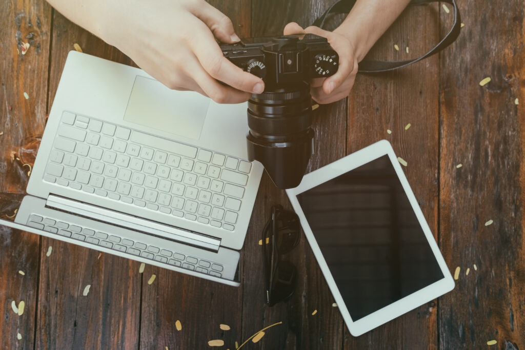 Hands holding a camera above a laptop and tablet on a wooden desk, symbolizing digital tools used in marketing vs PR for photographers to build clients and credibility.