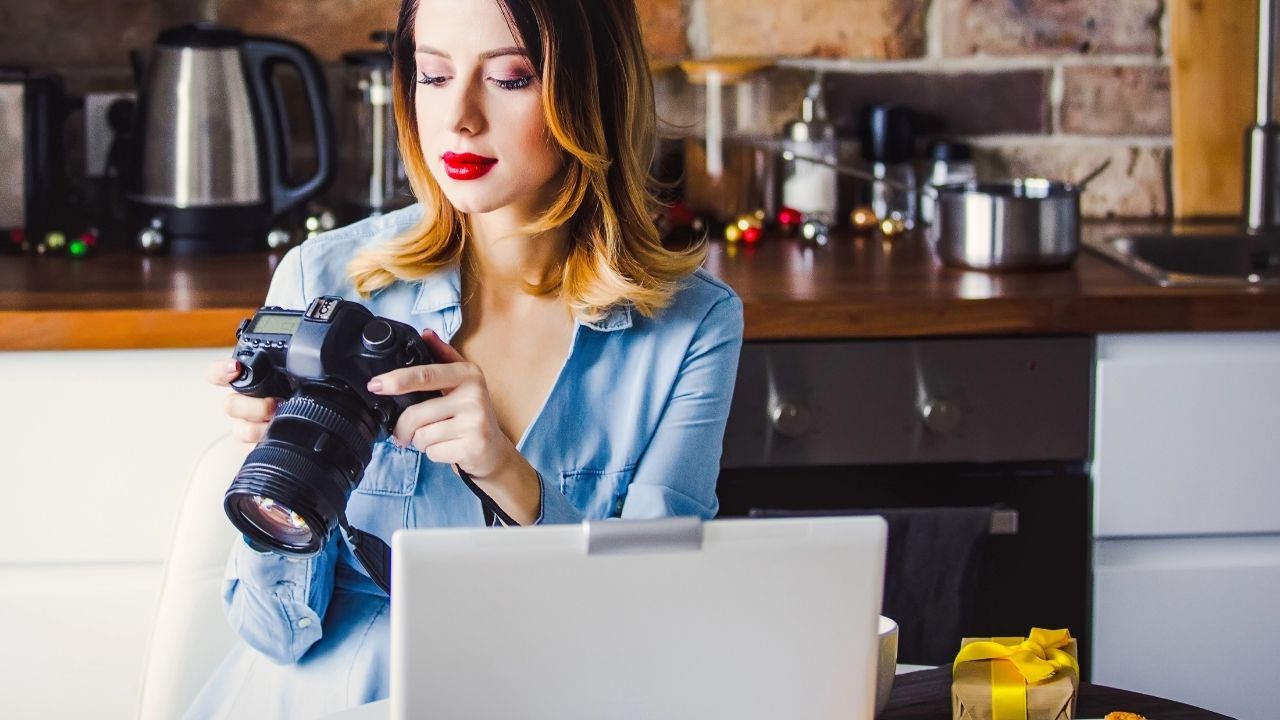 Female photographer reviewing her camera settings while working on a laptop in a home office, symbolizing marketing for photographers.