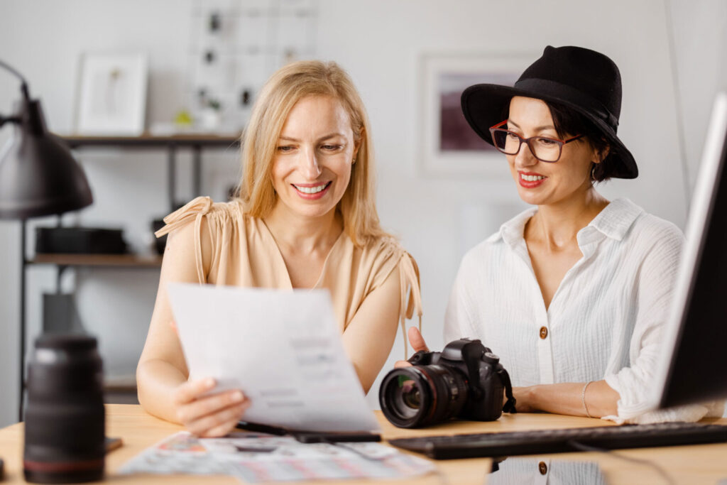 Two women reviewing photography business materials with a camera on the desk, representing collaboration and planning for Photography Black Friday Deals.