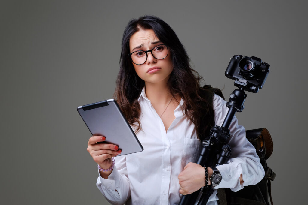 A confused female photographer holding a tablet and camera on a tripod, symbolizing the challenge of choosing a photography marketing agency to scale a studio.